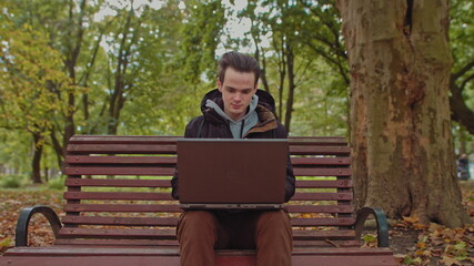 Fototapeta premium Medium shot of handsome young man working on laptop computer sitting outdoors in park, then looks at the camera and shows OK.Work alone is far from the office,freelancer
