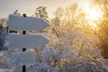 Naklejka premium Empty white track pointers, guidepost in sunlight against winter nature background. Directional arrow signs on wooden pole in snowy forest.