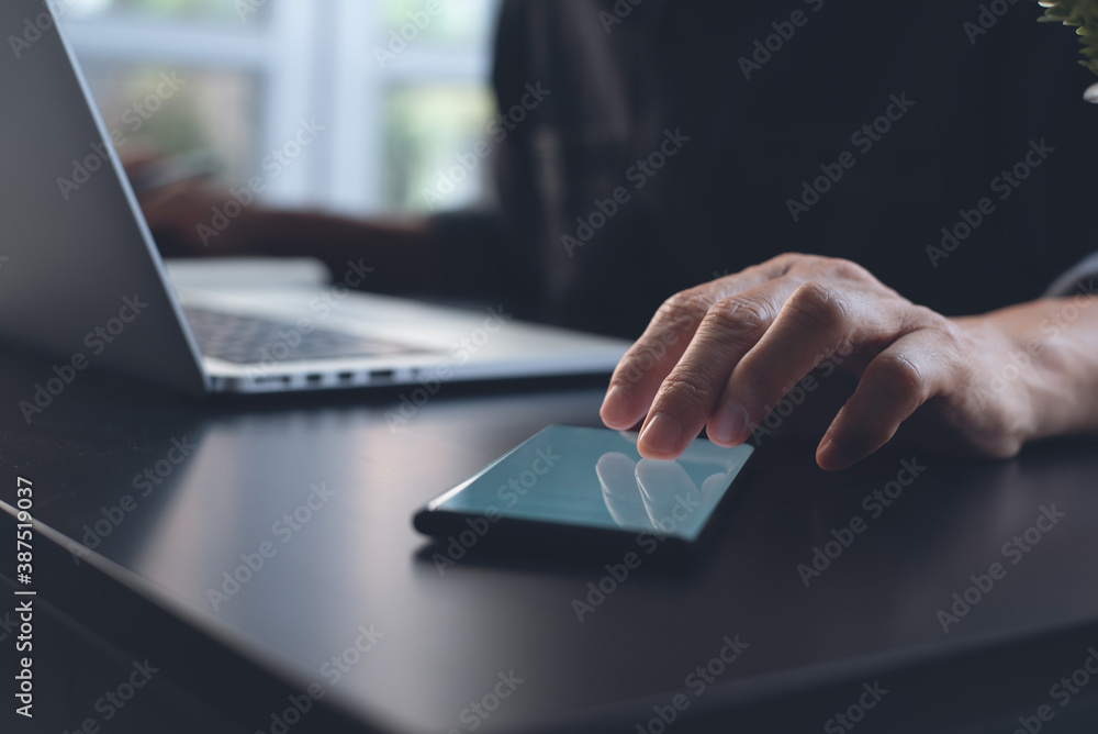 Poster man using mobile phone while working on laptop computer on wooden table in office