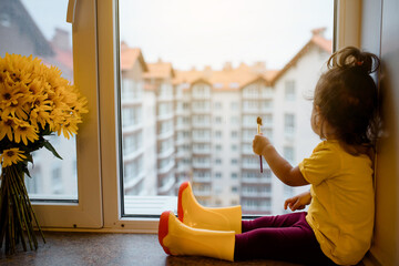 2 year child girl in a yellow t-shirt and yellow rain boots is sitting on a window sill near a vase...