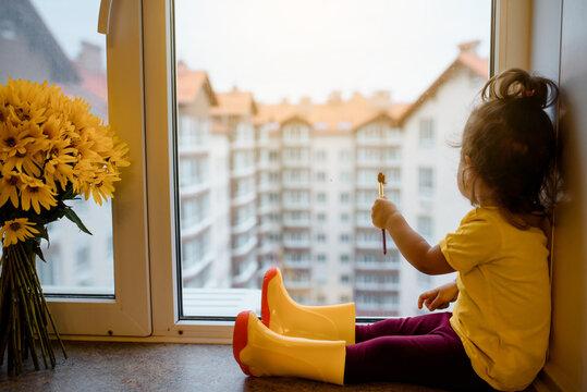 2 year child girl in a yellow t-shirt and yellow rain boots is sitting on a window sill near a vase with yellow flowers and paint with bruth on the window.