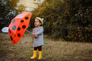 Happy smiling little child 2 years old girl in yellow rain boots is playing with  her red umbrella with polka dots outdoors.