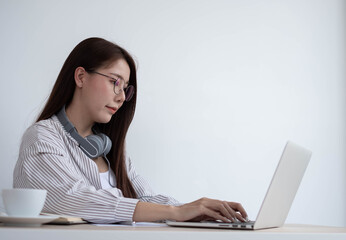 Young Asian women wear headphones using their laptop inside the office, working for online lessons or listening to webinars.