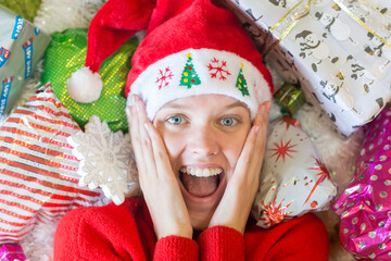 Christmas surprise! Excited amazed young woman lying next to many gifts wearing a Santa hat.