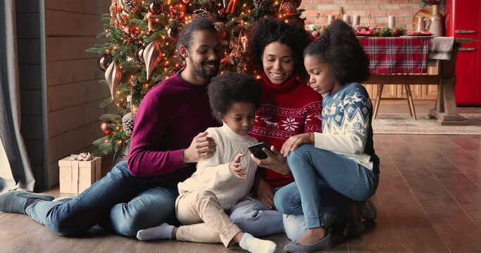 Full african family sit on warm floor in cozy modern living room spend winter holidays on internet using smartphone enjoy new trendy app taking selfie having fun together near decorated Christmas tree
