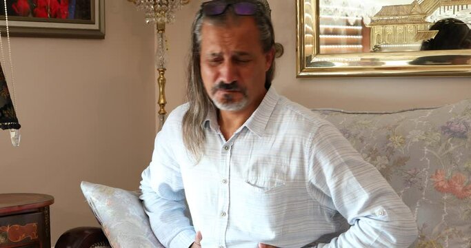 Portrait of a middle aged man sitting on sofa and having strong stomach ache  hands on abdomen