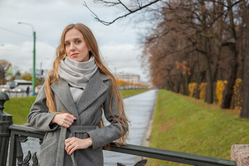 Fototapeta premium Portrait of a beautiful young woman on the background of the canal and autumn park. Attractive woman in a gray coat and scarf. Fall season. Autumn mood.