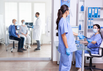 Fototapeta premium Medical nurse holding x-ray in hospital hallway during coronavirus outbreak wearing face mask . Assistant working on reception computer. Doctor dicussing with senior couple