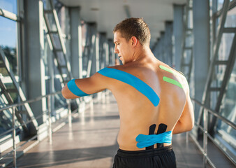 Fototapeta na wymiar Back view of muscularman wearing black sports outfit, looking aside. Young male athlete posing indoors, colorful kinesiology taping on naked back, futuristic interior. Sports rehabilitation concept.