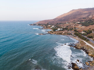 Aerial view of open Aegean sea at sunset, waves breaking with white foam on sand beach and stony coast, cloudy sky, Crete, Greece. Summer vacation. Mountainous landscape and seascape of Greek island.