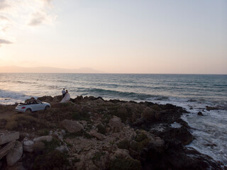 Bride in white fluffy dress and groom in tuxedo hugging on rocky coast, shore near the sea at sunset, white cabriolet car stand near, Crete, Greece. Wedding love story. Aerial.