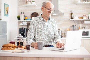 Senior man smiling and using laptop in the kitchen. Daily life of senior man in kitchen during breakfast using laptop holding a cup of coffee. Elderly retired person working from home, telecommuting