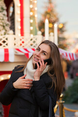 Young woman talking on a mobile phone walking at Christmas Market. Beautiful girl, warmly dressed enjoys Merry Christmas and Happy Holidays. Happy New Year. Christmas Eve