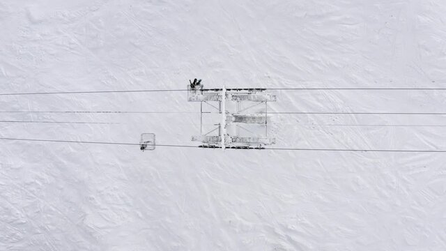 Overhead view from drone cabin chairlift moving on cable way in ski resort on snow background. Ski lift transporting skiers and snowboarders on winter slope at mountain resort. Ski elevator top view