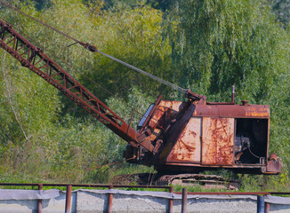 Fototapeta premium An old and rusty idle excavator is photographed standing on the bank of the river against the background of a green tree