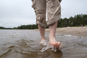 Barefoot male legs in rolled up pants walk in the water along the sandy shore, against the backdrop of a lake and trees. Lifestyle.