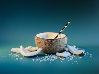 Half of coconut and pices of broken coco nut around it at dark background of teal and viridian colors. Opened cocos with organic vegetarian milk and coctail straw.