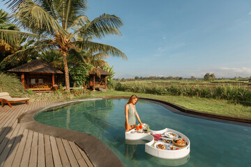 Obraz premium Girl relaxing and eating floating breakfast in jungle pool on luxury villa io Bali island
