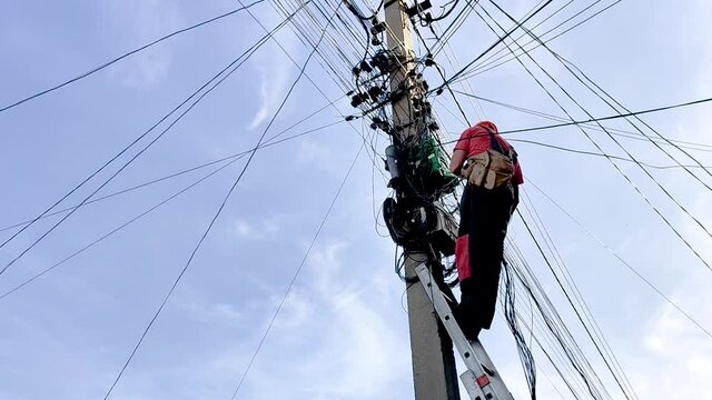 An electrician with a helmet and a safety belt standing on a ladder leaning against an electrical support among many tangled wires, cables and other communications.