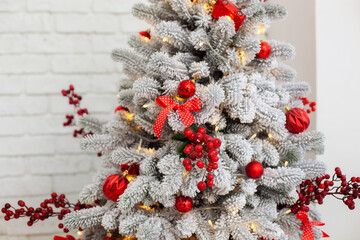 Xmas snow tree decorated with toys, branches of red berries, balls and lights garlands in home. Closeup view of beautiful decorated Christmas tree in living room. Christmas decorations concept. 