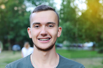 The young man is sincerely smiling and it is clear that he has ceramic braces on his teeth to align teeth. Guy on a background of nature with braces. Orthodontic concept