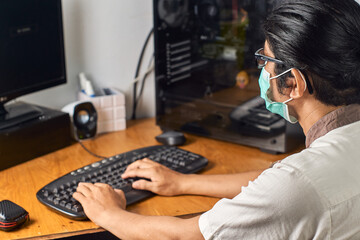Young asian man use medical mask and looking at computer monitor during working day from home. communicating in social network, searching information online at pandemic situation.