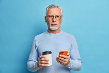 Senior gentleman uses modern smartphone and has coffee uses new technologies looks seriously at camera wears eyeglasses poses against blue background. Mature entrepreneur has break surfs internet