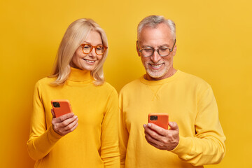 Photo of elderly grandmother and grandfather view photos together on smartphone devices watch interesting funny video online dressed in casual yellow turtlenecks pose indoor. Age and technology