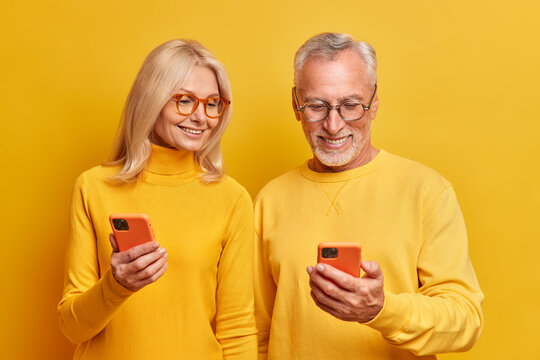 Photo of elderly grandmother and grandfather view photos together on smartphone devices watch interesting funny video online dressed in casual yellow turtlenecks pose indoor. Age and technology