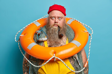Stunned bearded man stares bugged eyes poses with fishing net lifebuoy and smokes pipe spends time...