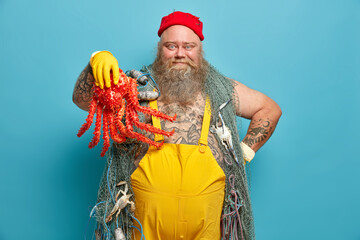 Handsome cheerful bearded boatswain holds big red octopus dressed in yellow overalls happy to catch sea creature carries fishing net poses against blue background. Angling at sea marine travel
