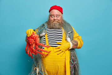 Cheerful bearded fisherman keeps hand on belly happy to catch big octopus enjoys marine adventure and voyage looks surprisingly at camera poses indoor against blue background. Middle aged sailor