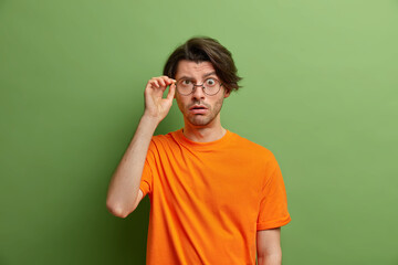 Half length shot of surprised guy looks with wonder through spectacles holds breath dressed in casual wear hears astonishing news isolated over green background. Human reactions and emotions concept