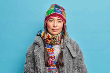 Serious Scandinavian woman with pigtails looks calmly at camera dressed in warm winter clothing poses over blue background. Eskimo girl wears fur coat and knitted hat for cold season wrapped by scarf - obrazy, fototapety, plakaty