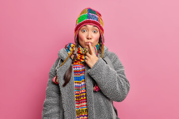 Surprised teenage girl keeps index finger on lips and looks stunned at camera dressed in grey fur coat and knitted hat isolated on pink background. Nothern woman in traditional warm clothes.