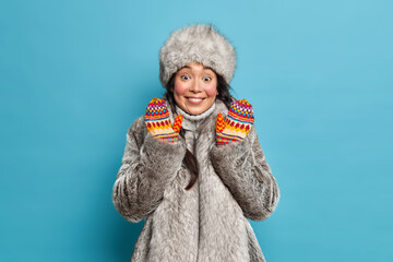 Winter time cold season concept. Cheerful Eskimo woman dressed in fur hat coat and gloves smiles happily keeps hands raised lives at north isolated on blue background. Arctic native inhabitant - obrazy, fototapety, plakaty