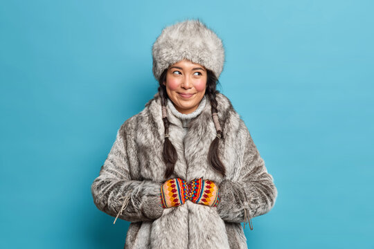 Pleased smiling eskimos woman dressed in traditional clothing adapted to extreme cold lives on nothern pole isolated over blue background. Female inhabitant of Alaska wears fut coat and hat.
