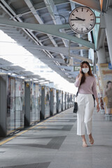 Asian business woman in casual clothes wearing face mask talking on mobile phone. She is waiting for the train to go to work on the platform station. New normal lifestyle in city concept.