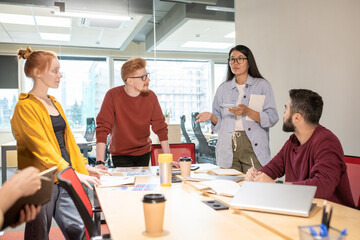 Young Asian female broker standing by table and explaining her point of view