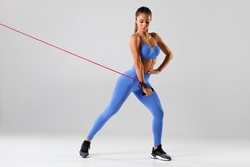 Fototapeta premium Fitness woman exercises with resistance band on gray background. Athletic girl working out