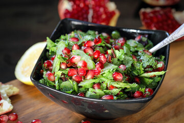 Tabbouleh with pomegranate. Green salad of parsley and herring with large pomegranate grains, in a black plate on a dark background and a wooden board.