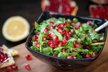 Tabbouleh with pomegranate. Green salad of parsley and herring with large pomegranate grains, in a black plate on a dark background and a wooden board.