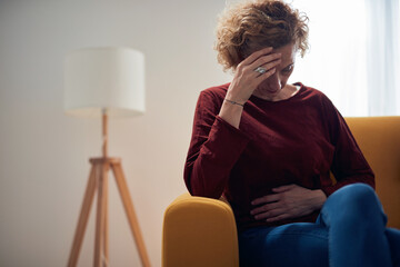 Woman with stomach pain and headache problem sitting on a couch at home.