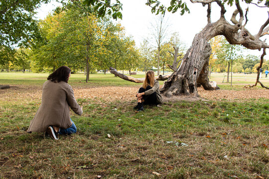 Male photographer taking photos of a blond model that is sitting on the grass next to a tree at Hyde Park, London, United Kingdom