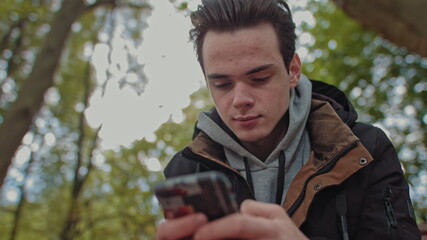 Teenager Looks at the phone, dials a message on a touch screen phone. Shooting close up of a smile on his face. The guy is sitting in the park on a bench. Corresponds with friends