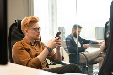 Young serious businessman in eyeglasses texting in smartphone in armchair