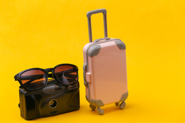 Travel concept. Mini pink plastic travel suitcase with camera, sunglasses on yellow background. Minimal style