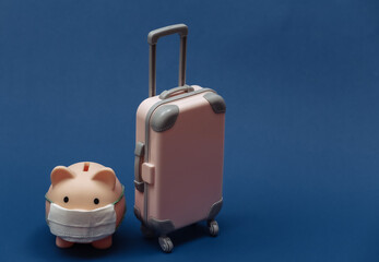 Travel during covid-19. Mini plastic travel suitcase and piggy bank with face mask on classic blue background.