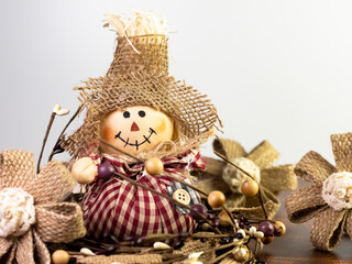 Mini Scarecrow with a Burlap Hat and red plaid outfit Surrounded by Burlap Flowers on a wooden surface with a white wall.  Fall autumn decor for the season.
