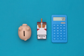 Pack of cigarettes and calculator, piggy bank on blue background. Smoking costs